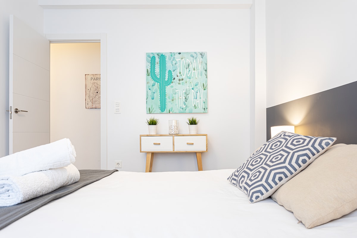 The bedroom features a spacious bed adorned with neutral-toned pillows and neatly arranged towels. A colorful wall art depicting cacti is displayed above a modern bedside table, which holds small green plants. Soft lighting enhances the bright, minimalist decor of the room.