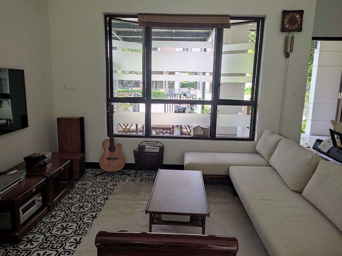 The living area features a large, light-colored sectional sofa positioned around a simple wooden coffee table. A wooden guitar leans against the wall, while a television is mounted opposite. Natural light enters through large windows overlooking a covered outdoor seating area.