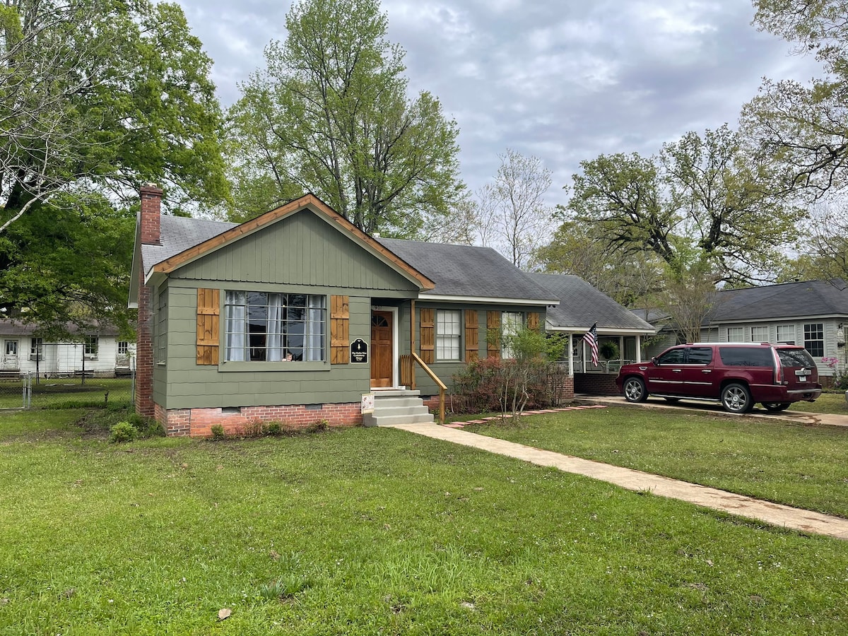 The front view of a charming house features a green exterior with wooden shutters and a welcoming porch. A manicured lawn surrounds the property, complemented by tall trees and nearby residential homes. A driveway leads to a parked vehicle, providing easy access.
