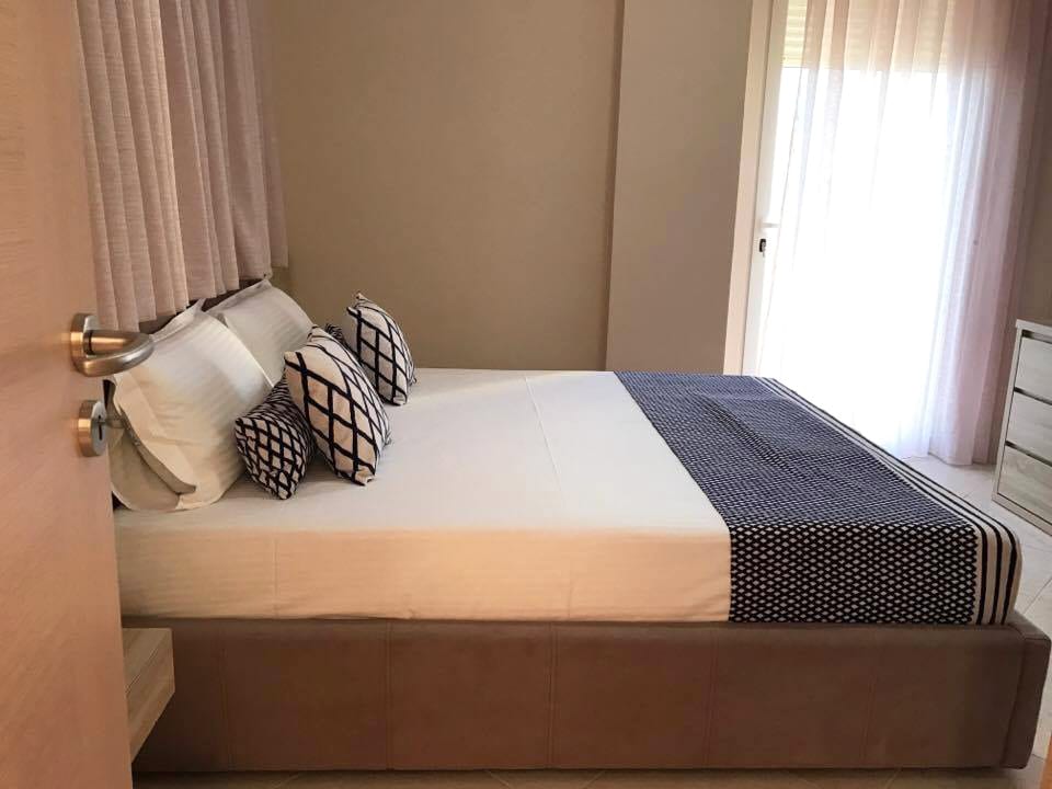 A spacious bedroom features a large bed with crisp white linens and decorative pillows. Natural light filters through sheer curtains, creating a bright and airy environment. A low wooden bedside table is positioned next to the bed, complementing the minimalistic decor.