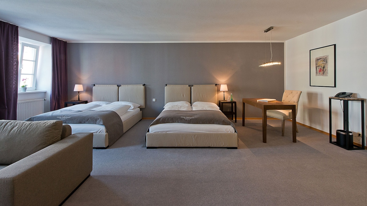 A spacious living and sleeping area is presented, featuring two beds and a comfortable sofa. Minimalist decor is complemented by soft lighting, while a decorative table and chairs provide a functional space for dining or working. Windows are adorned with dark curtains.