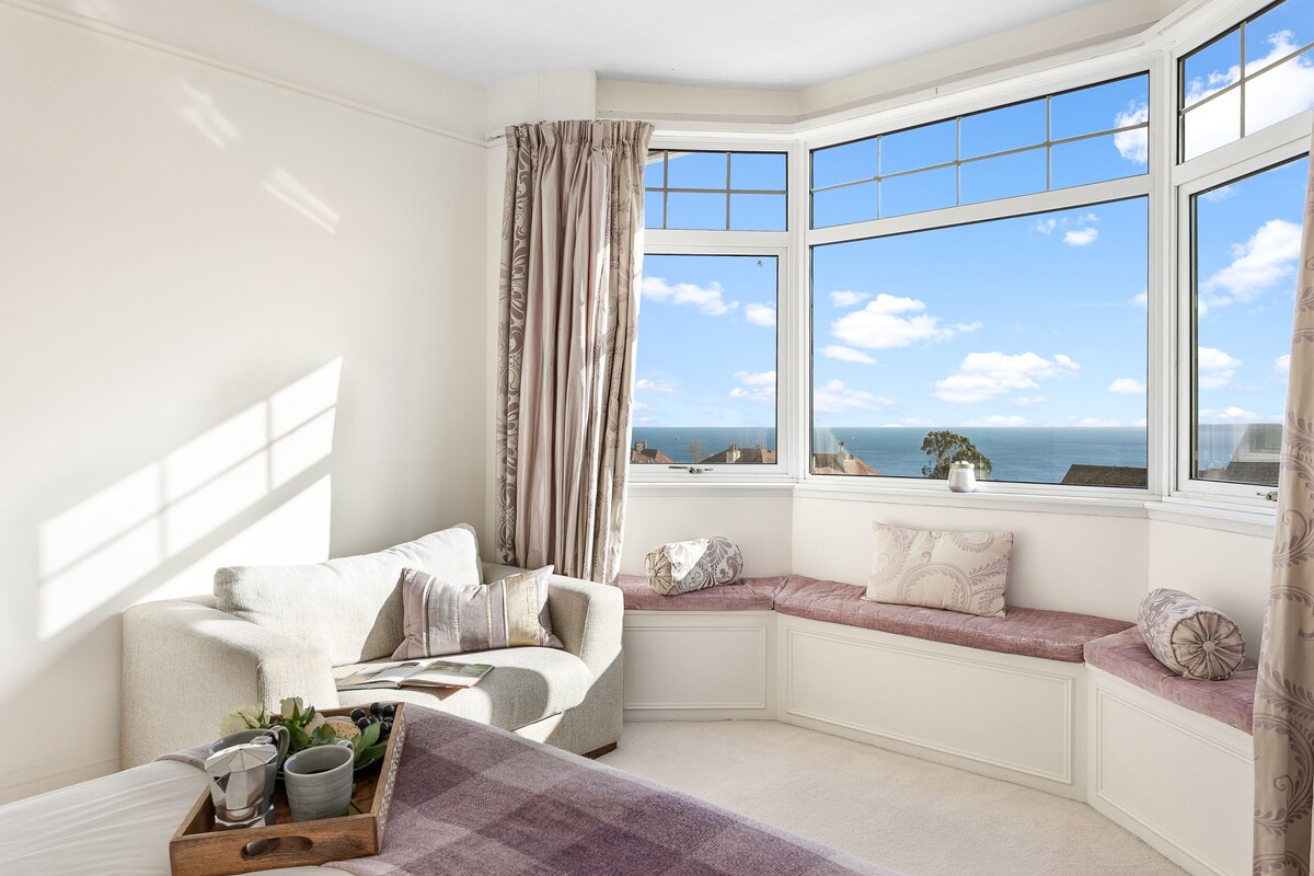 A bright corner of a room features a bay window with panoramic views of the sea. Comfortable seating is arranged along the window ledge, complemented by cushions. A cozy throw is draped over the bed, while natural light fills the space, enhancing its airy feel.