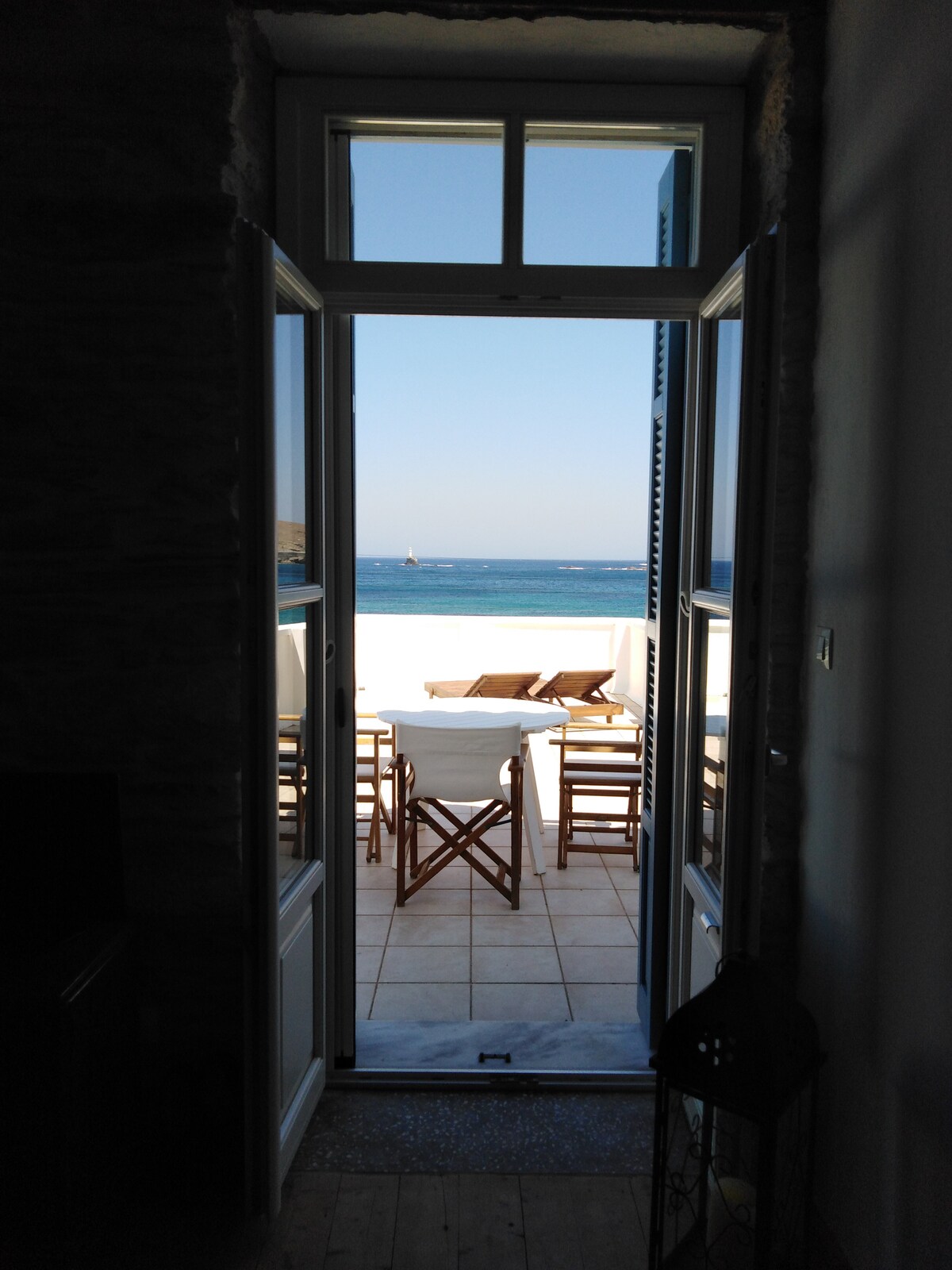A view through an open door shows a bright outdoor terrace with wooden furniture and sun loungers. The sea is visible in the background, reflecting sunlight, creating a serene coastal atmosphere.