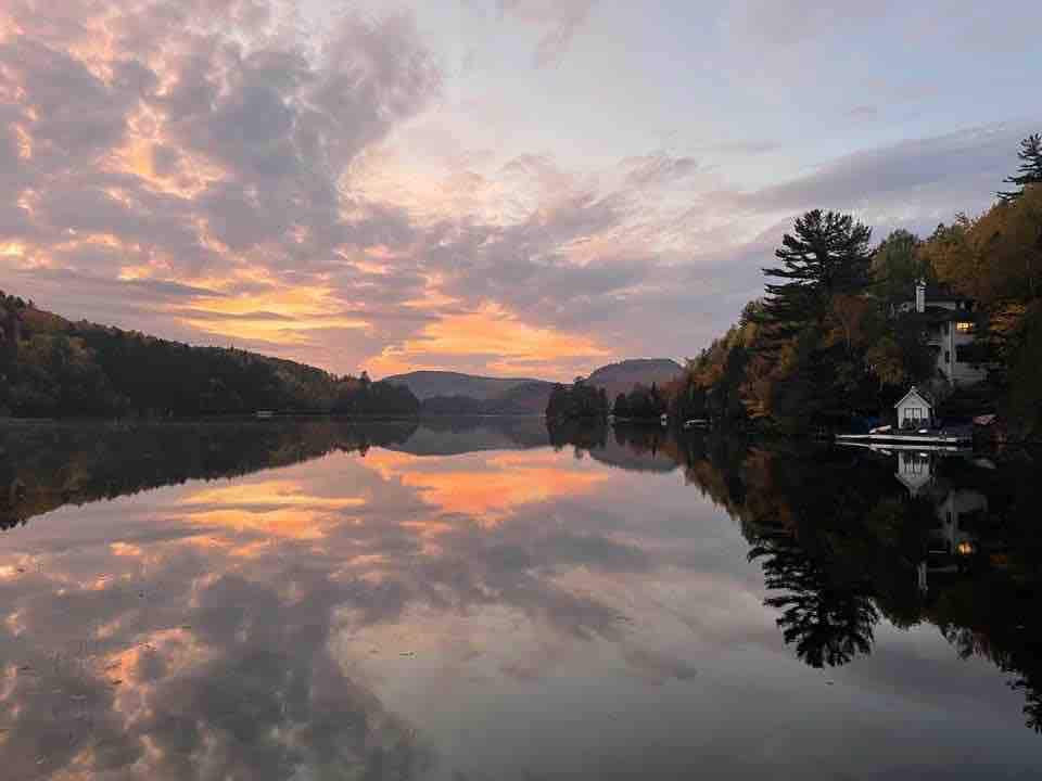A serene lake is depicted during sunset, reflecting a vibrant sky filled with soft clouds in shades of orange and pink. The surrounding landscape features lush trees and mountains, creating a tranquil atmosphere. The water's surface appears calm and glassy, enhancing the picturesque scene.