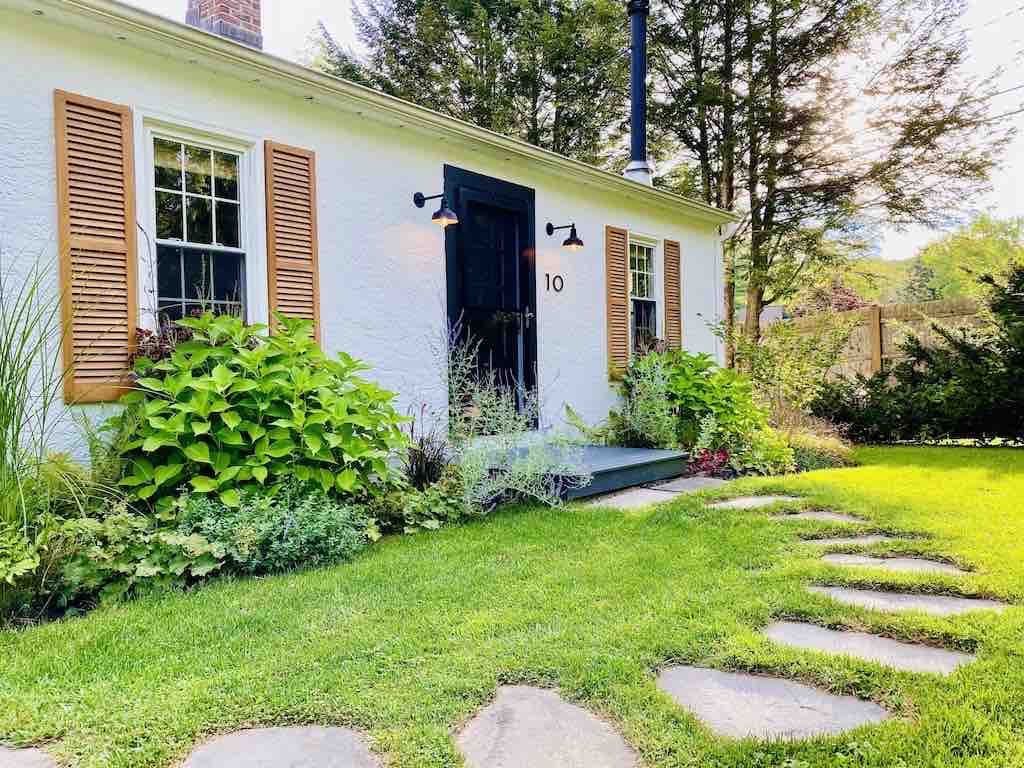 The entrance of a charming cottage features a clean, white stucco facade accented by wooden shutters. A welcoming black door stands at the center, surrounded by lush greenery and colorful plants. A stone pathway leads to the entrance, enhancing the cozy outdoor atmosphere.