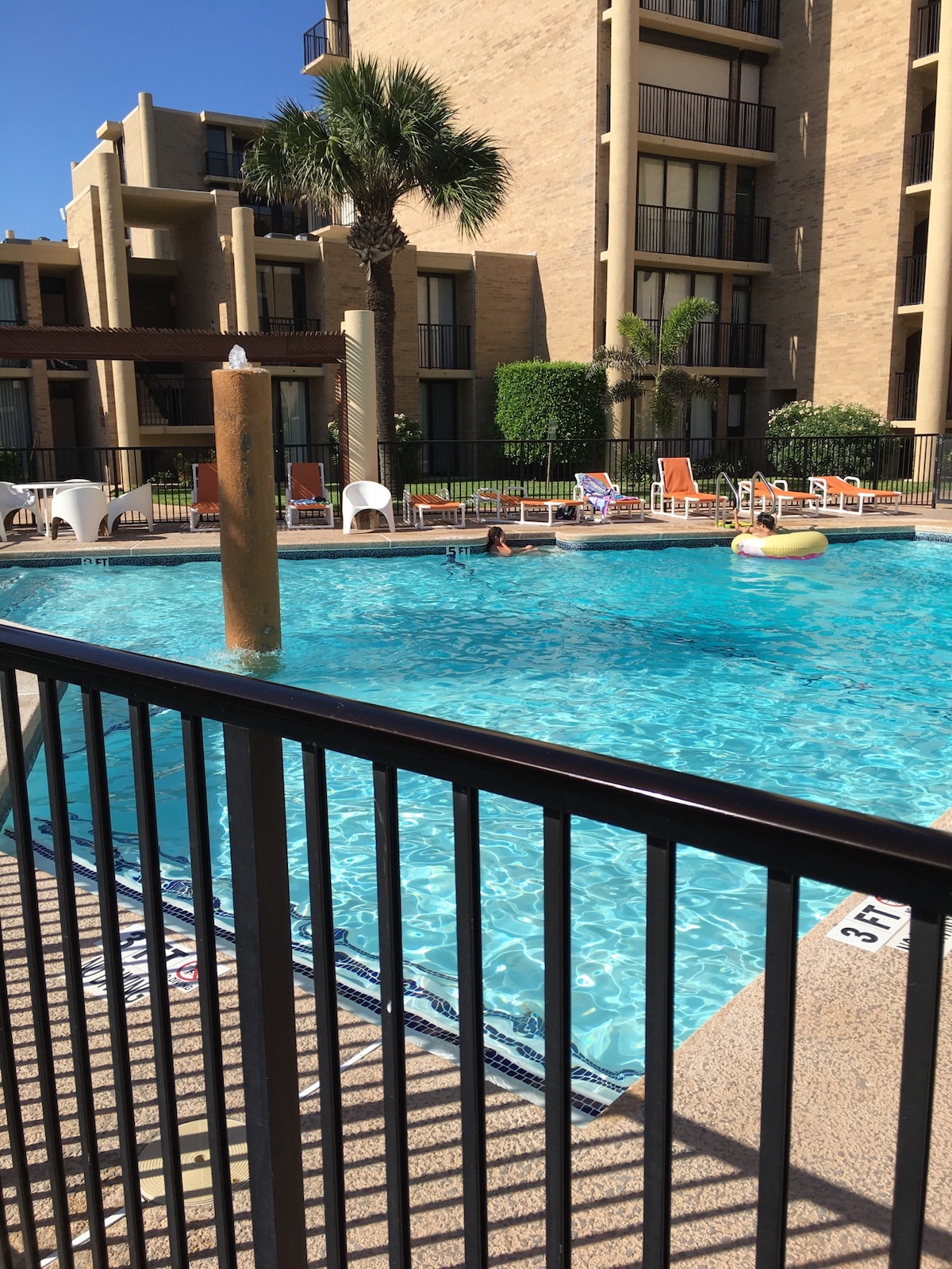 A spacious outdoor pool area features crystal-clear water under a bright sky. Palm trees provide shade, while lounge chairs are neatly arranged around the pool. A person enjoys the water with a floating device, and a fence ensures safety around the poolside.