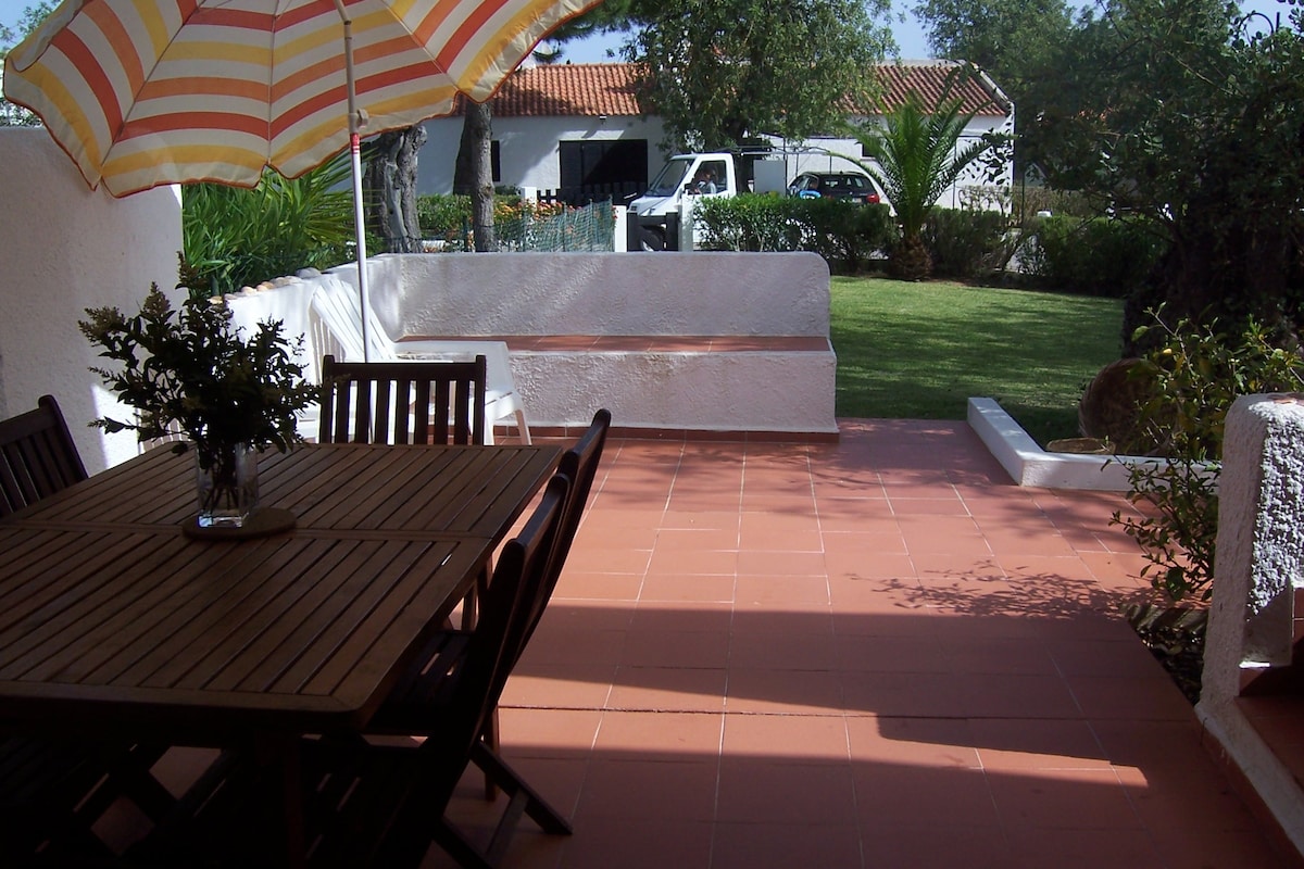 An inviting outdoor terrace features a wooden dining table accompanied by several matching chairs. A large striped umbrella provides shade. The patio is bordered by neatly trimmed grass and surrounded by greenery, creating a serene outdoor space for meals or relaxation.