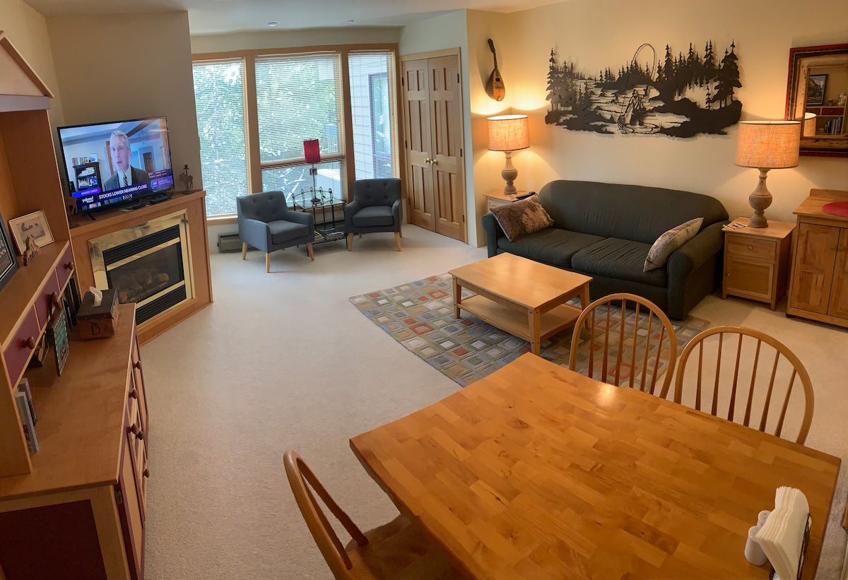 A welcoming living space features a dark couch facing a television. Light wooden furniture includes a dining table with four chairs and a coffee table. A carpet with a geometric design adds texture. Natural light filters through large windows, providing a view of the outside.