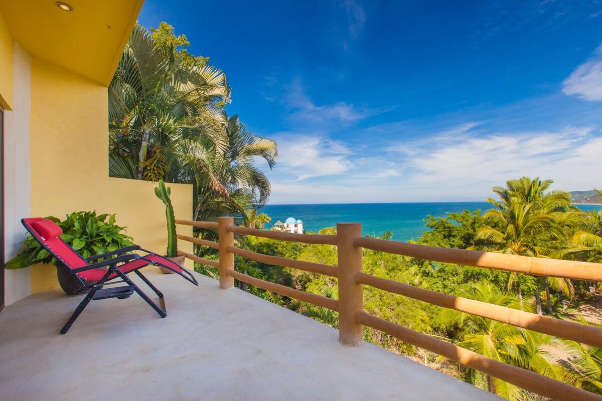 A balcony is presented with a view of the ocean and coastline, surrounded by lush greenery. A single red lounge chair rests against the railing, inviting relaxation. The sky is bright blue with scattered clouds, enhancing the sense of openness and tranquility.