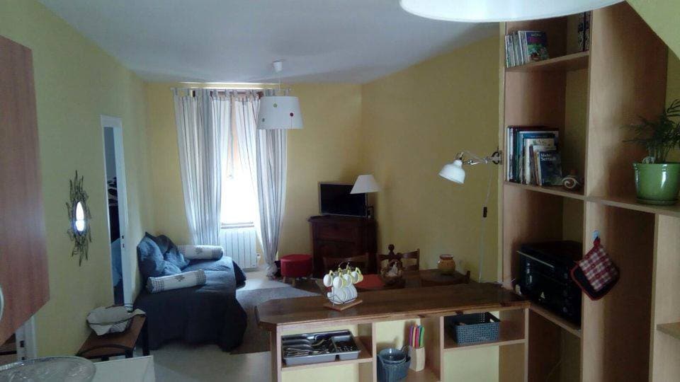 A bright living area features yellow walls and a comfortable seating arrangement. A small television is positioned on a wooden stand, complemented by a stylish light fixture above. A bookshelf holds various items, and natural light streams through sheer curtains.