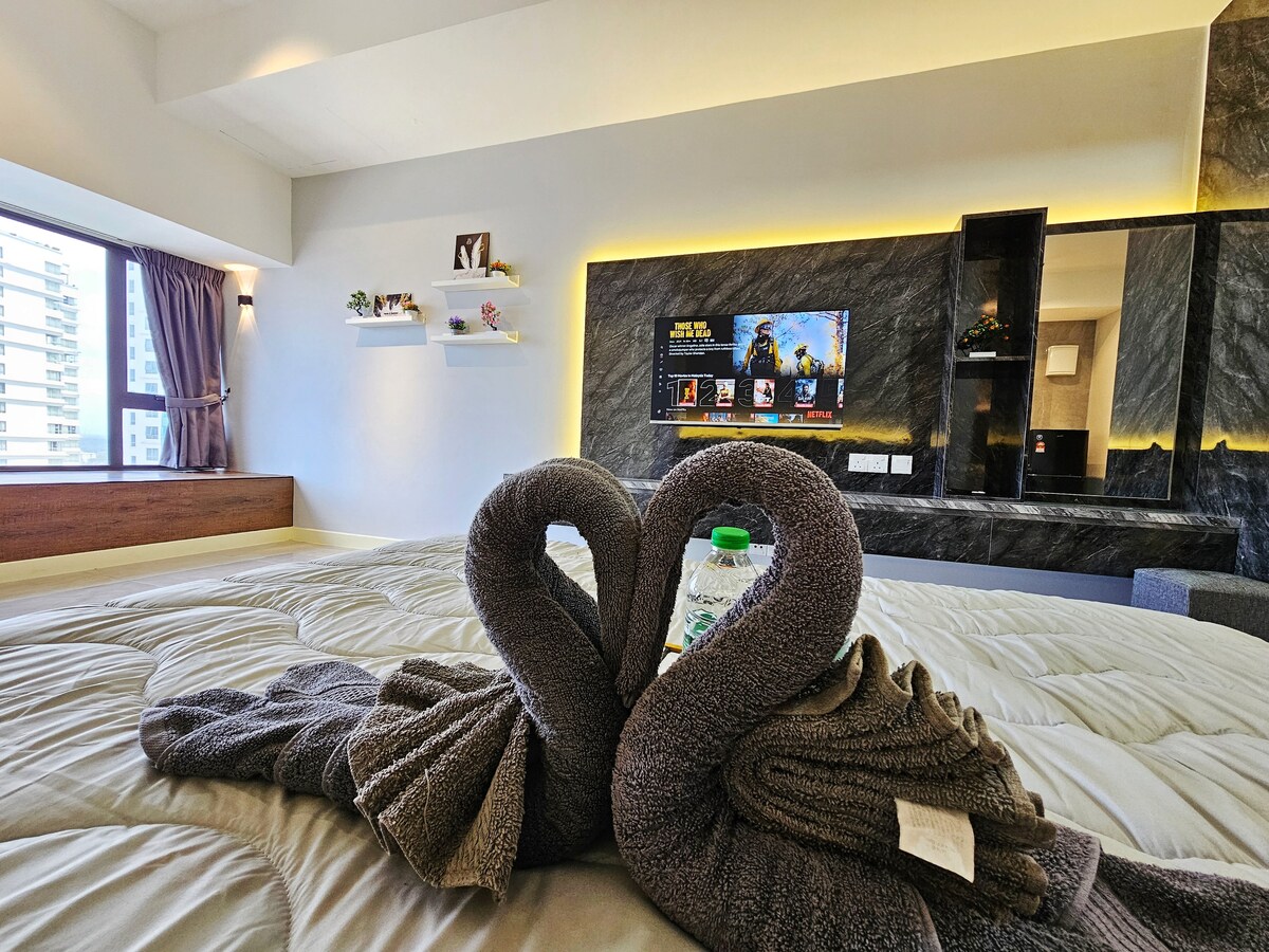 A spacious bed is adorned with neatly arranged towels shaped like swans. A television mounted on the wall is visible, alongside shelves displaying decorative items. Natural light filters through a large window dressed with curtains, illuminating the modern interior design.