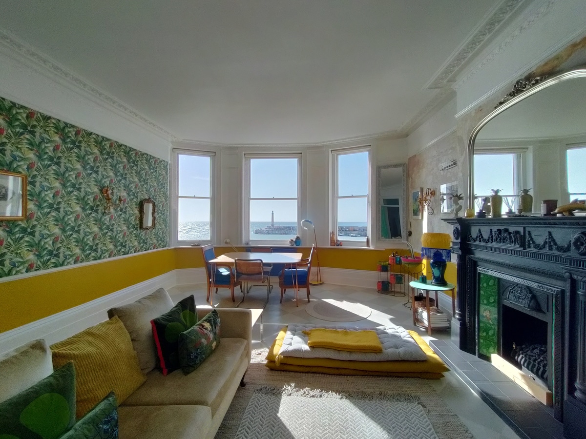 A spacious living room is featured with large windows offering sea views. Vintage furniture and a decorative fireplace complement bright tropical wallpaper. A casual seating area with cushions is visible, while a dining area accommodates a table and chairs, enhanced by natural light.