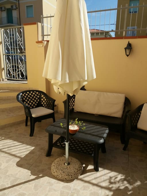 Cortile interno casa con una poltrona grande e due sedie in plastica. Ombrello con cassa acustica (con bluetooth incorporato).