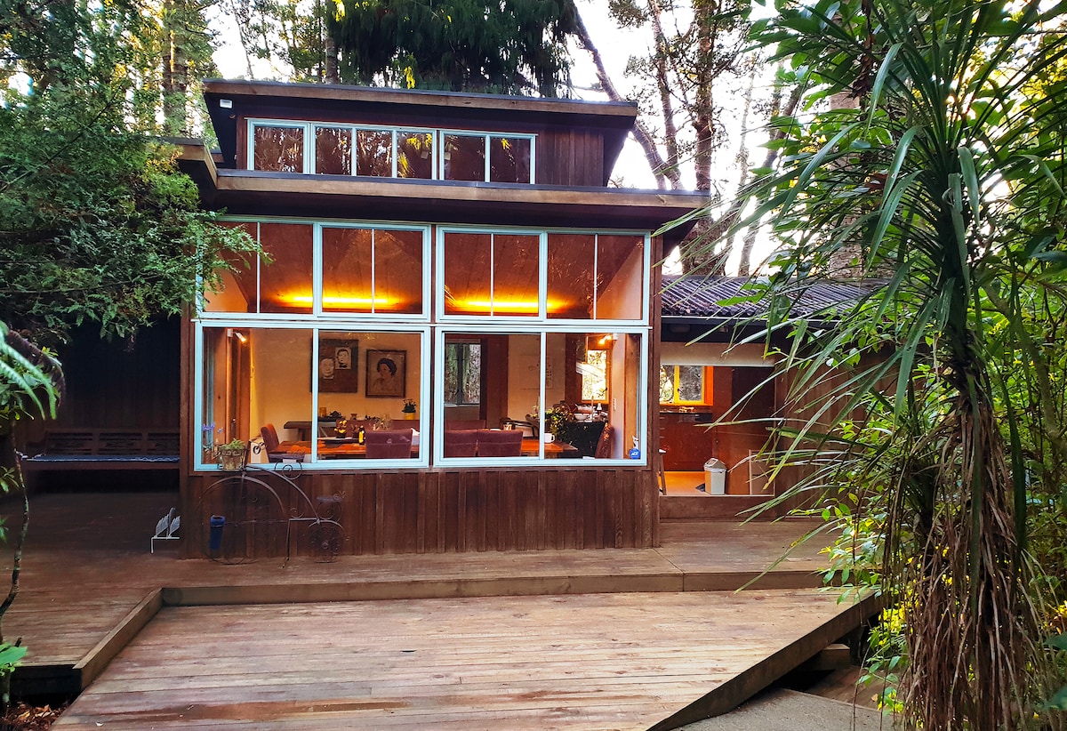 A spacious wooden exterior is surrounded by lush greenery, featuring large glass windows that invite natural light into an open living area. The deck offers a smooth wooden surface leading to a cozy lounge space visible through the windows.
