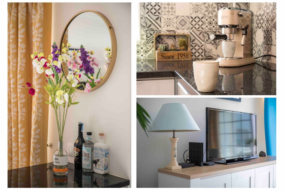 A round mirror is positioned above a black countertop adorned with a vase of colorful flowers. Beside it, a coffee maker and a cup are arranged on the surface. A light blue lamp and a flat-screen television are placed on a wooden console, contributing to the modern decor.