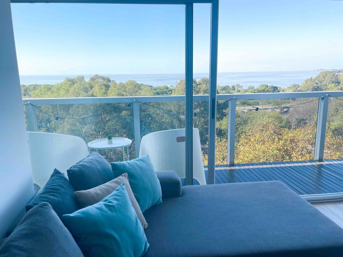 A spacious seating area features a comfortable gray sofa adorned with decorative pillows. Two light chairs and a small table are positioned on a balcony, offering a view of the lush greenery and expansive water in the background.