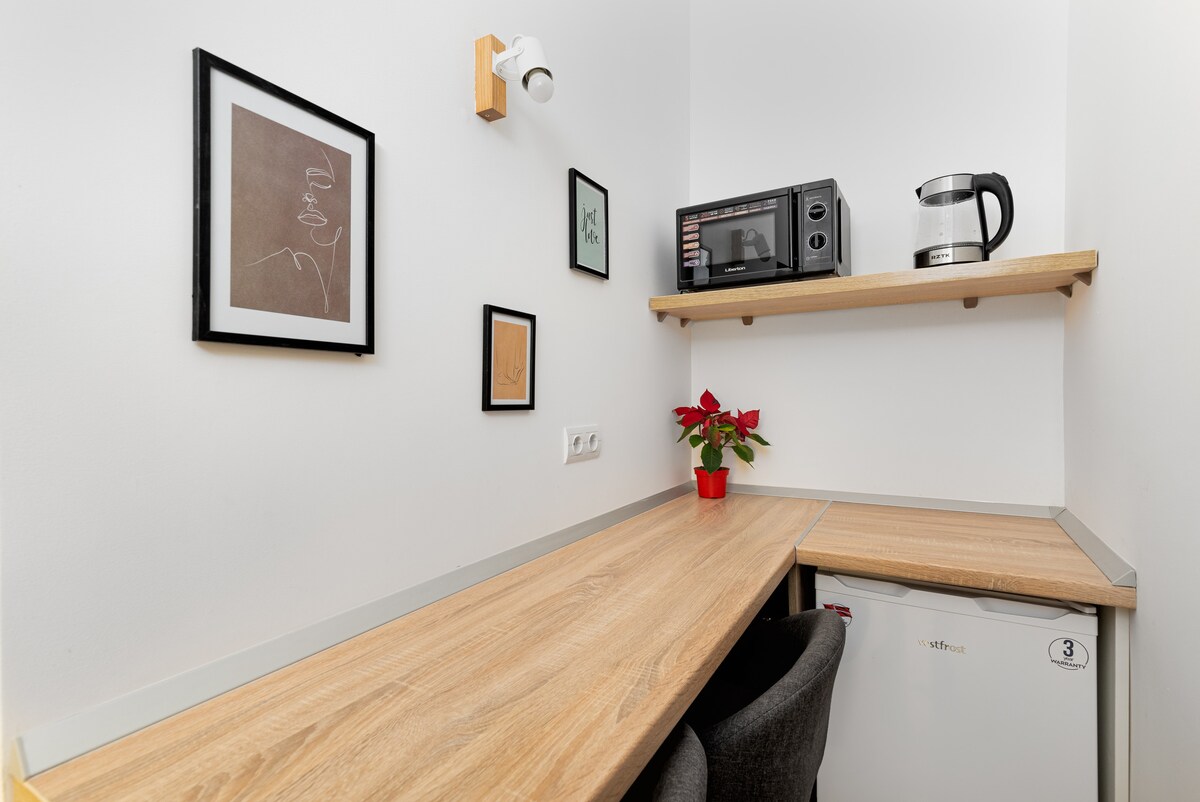A compact kitchen area features a wooden countertop with an integrated microwave and a kettle. A small refrigerator is positioned below, and a potted plant adds a touch of greenery. The walls display framed artwork, contributing to a pleasant and functional atmosphere.