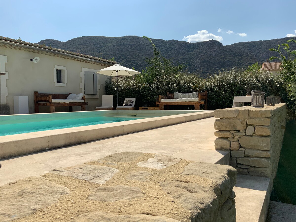 A tranquil outdoor space features a sunlit pool bordered by a stone wall. Wooden lounge furniture provides seating options under a white umbrella, surrounded by lush greenery. The backdrop showcases rolling hills under a clear blue sky.