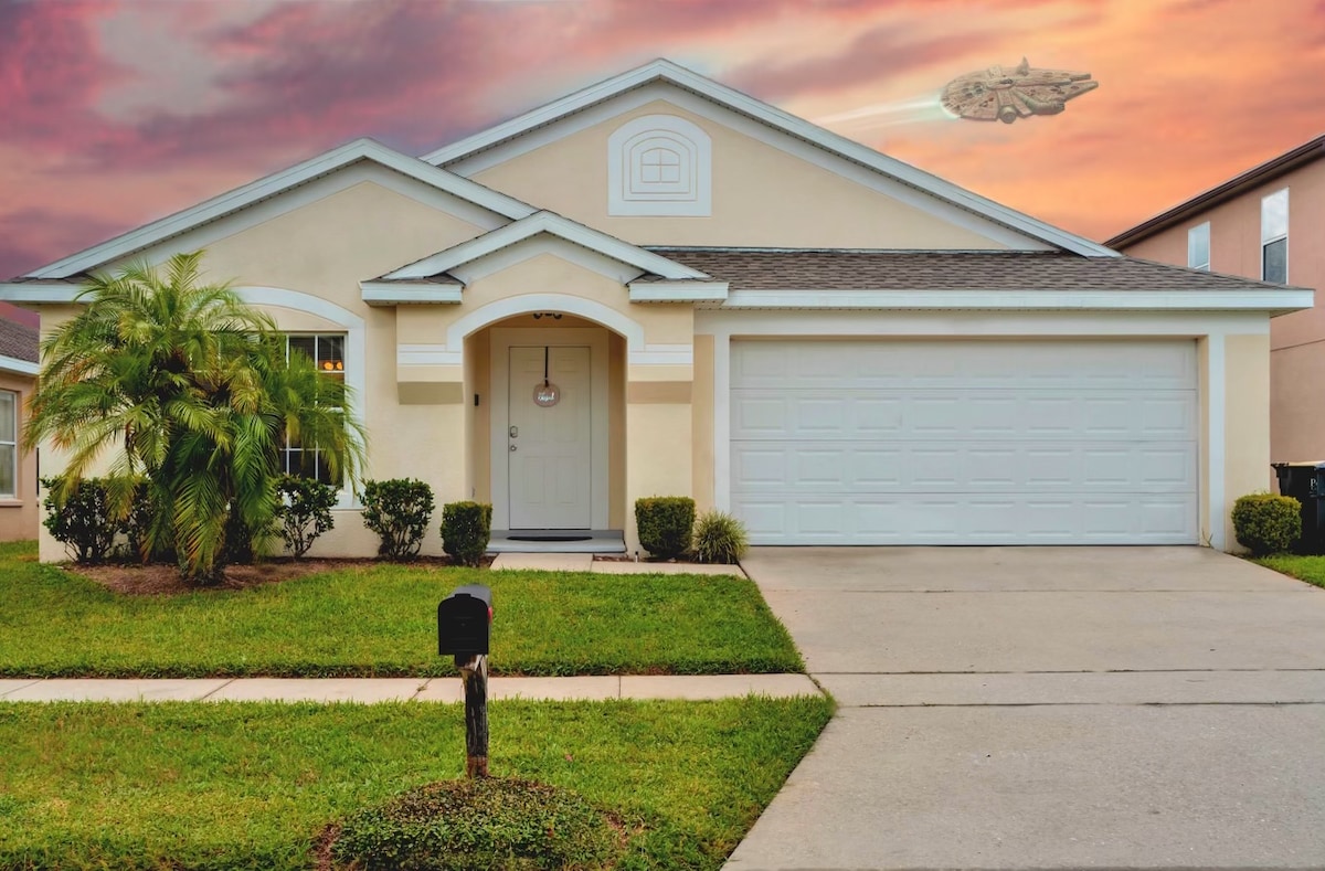 The exterior of a single-story ranch house is depicted, with a neatly manicured lawn and colorful bushes. A driveway leads to a two-car garage. A digital image of a spaceship floats above the house against a colorful sunset sky.