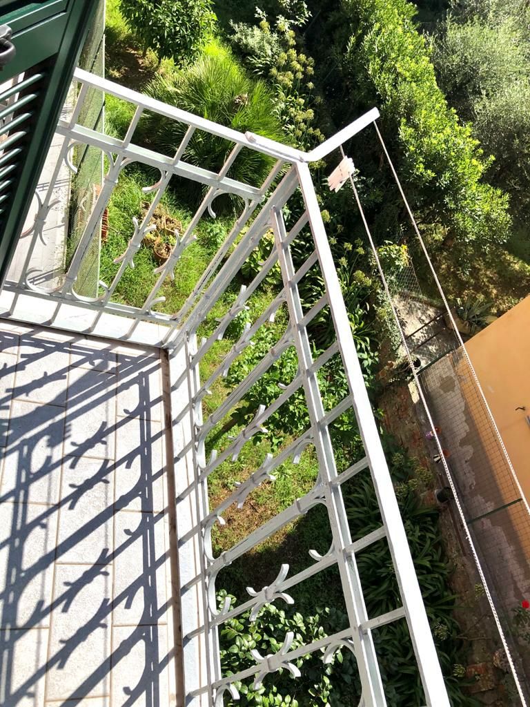 A balcony is shown with a wrought-iron railing overlooking a lush garden below. Sunlight filters through greenery, casting delicate shadows on the floor. The surrounding plants create a serene and private atmosphere, ideal for relaxation.