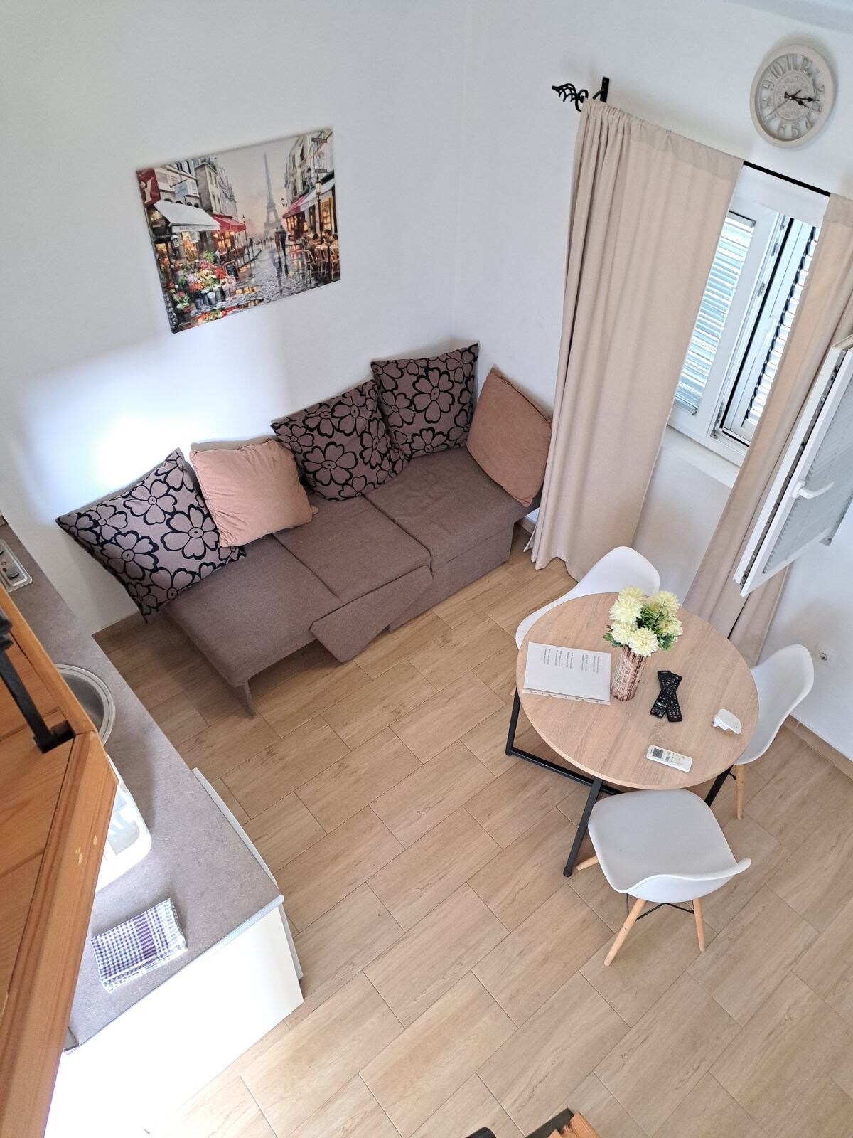 A view of the cozy living area is presented, with a modern sofa featuring decorative cushions arranged around a small dining table. Natural light enters through the window, which is dressed with light curtains. A framed artwork enhances the wall, contributing to the overall aesthetic.