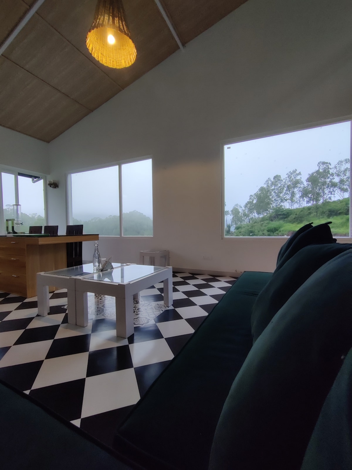 A modern living area is presented with large windows that invite natural light and offer views of the misty landscape. The space features a stylish checkered floor and a comfortable seating arrangement, complemented by a simple coffee table in the center.