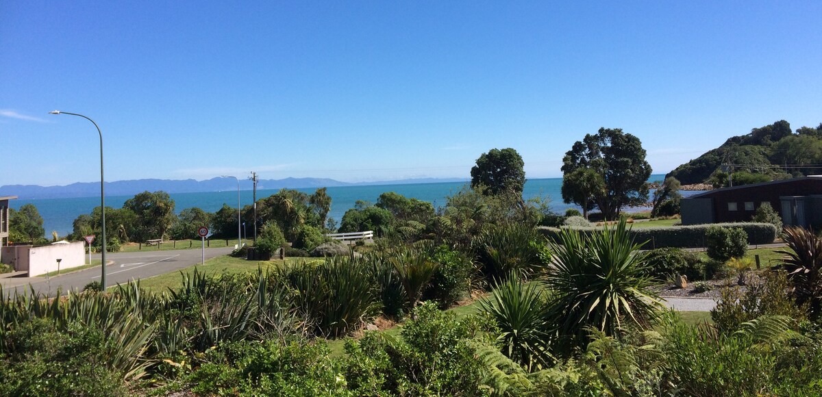 A scenic view showcases the tranquil ocean and distant mountains, framed by lush greenery and native plants in the foreground. A clear blue sky enhances the serene atmosphere, highlighting the peaceful coastal setting.