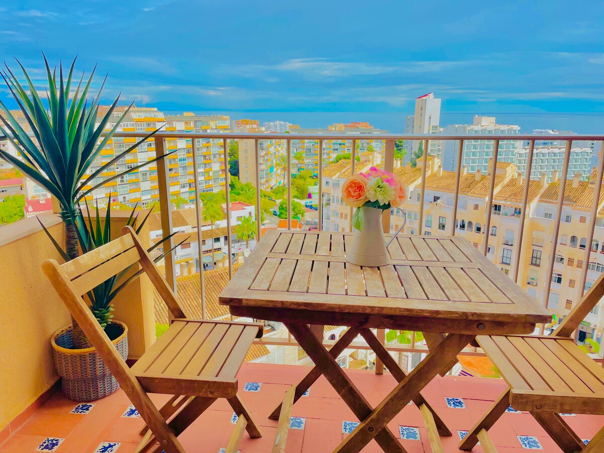 A small wooden table is set on a terrace, accompanied by two matching chairs. A vase with fresh flowers sits at the center. In the background, views of the ocean and surrounding buildings are visible under a clear blue sky.