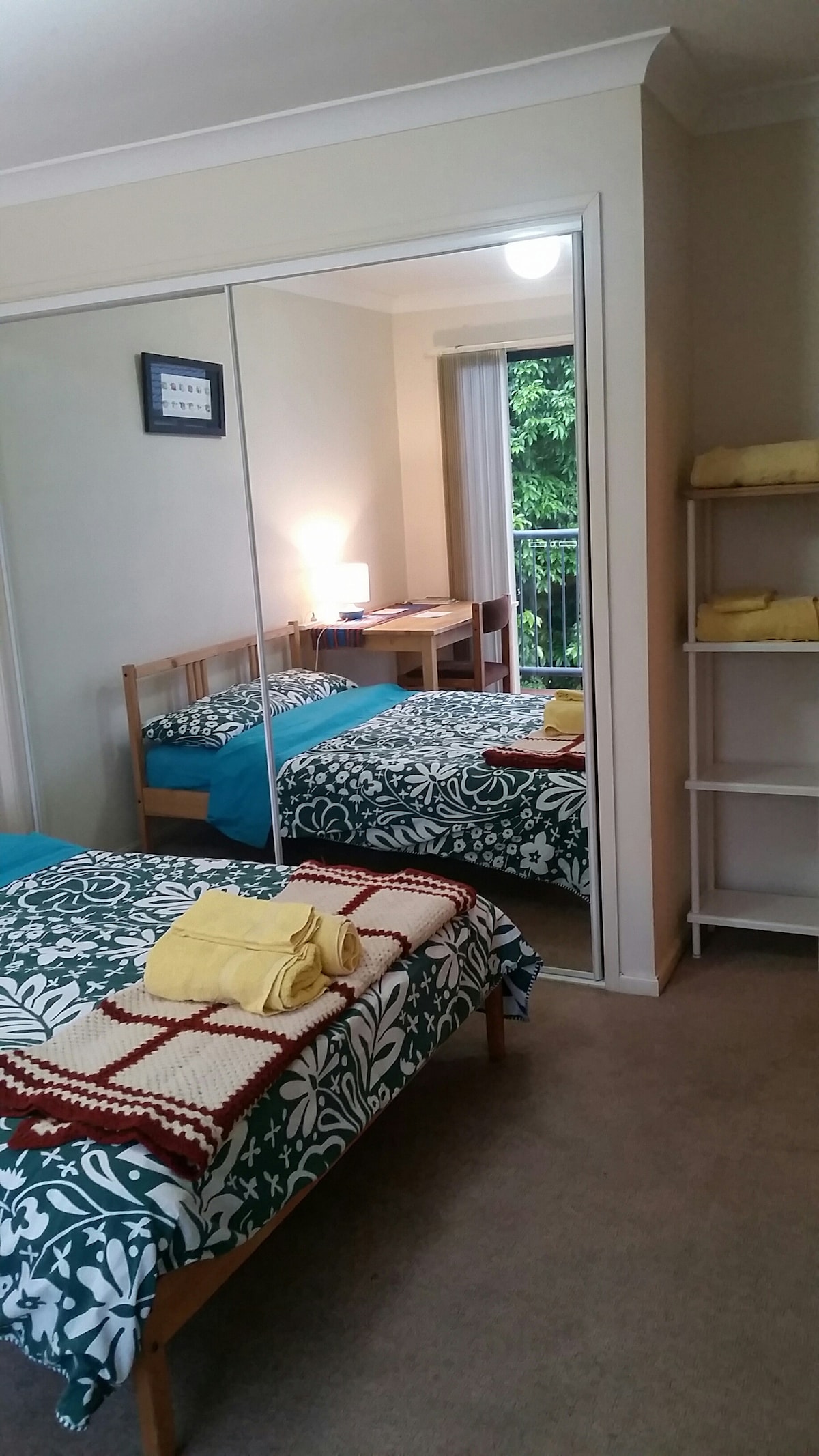 A roomy master bedroom is presented with a large mirror wardrobe and a comfortable bed adorned with a patterned duvet and neatly folded towels. A desk and light are visible, while a sliding door leads to a small deck area surrounded by greenery.
