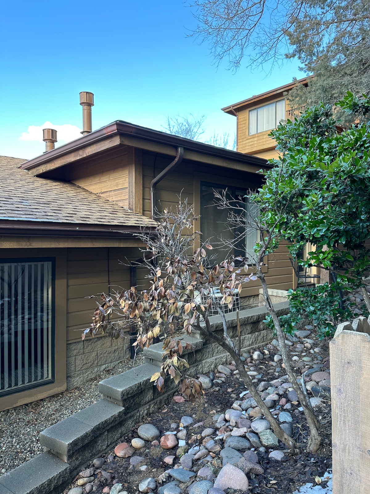 The exterior of the condo is framed by natural landscaping, featuring a pathway lined with smooth stones. A mix of trees and shrubs adds greenery around the entrance, while the building's wooden facade harmonizes with the environment. A clear blue sky serves as the backdrop.
