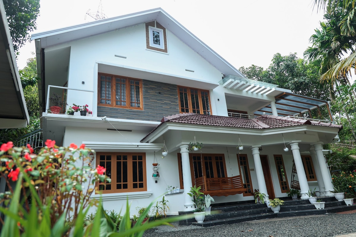 A modern two-story home is set amidst lush greenery, featuring a combination of wooden window accents and a light exterior. A covered porch with pillars welcomes guests, while planters enhance the entryway. The spacious balconies overlook the garden, providing an inviting atmosphere.