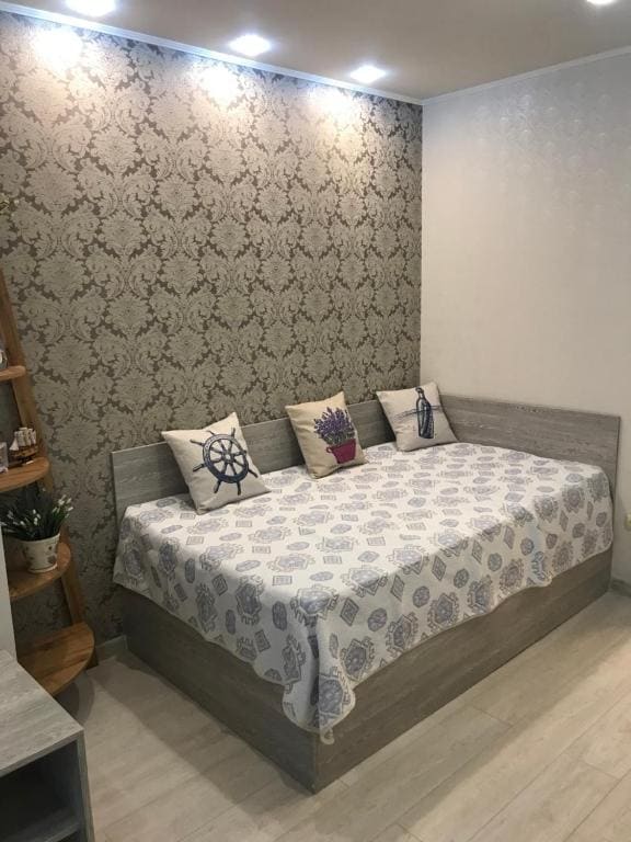 A lightly decorated sleeping area features a spacious bed with a patterned throw and decorative pillows. A subtle floral wallpaper complements the modern furnishings, including a small shelf displaying plants. Soft lighting is provided by recessed ceiling fixtures.