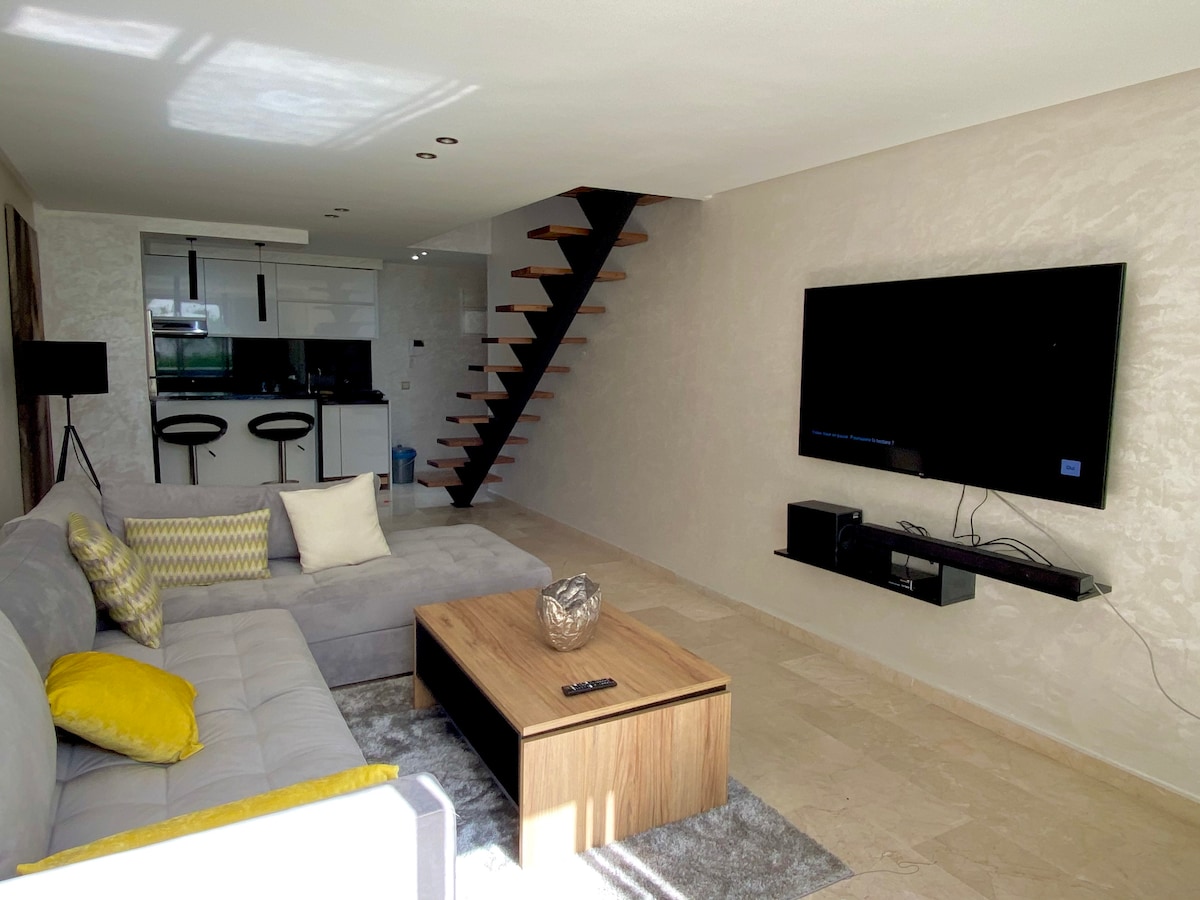 A comfortable living area features a grey sectional sofa adorned with yellow cushions. A wooden coffee table stands in the center, while a wall-mounted television is positioned opposite the seating. Light filters through the space, highlighting a staircase that leads to an upper level.