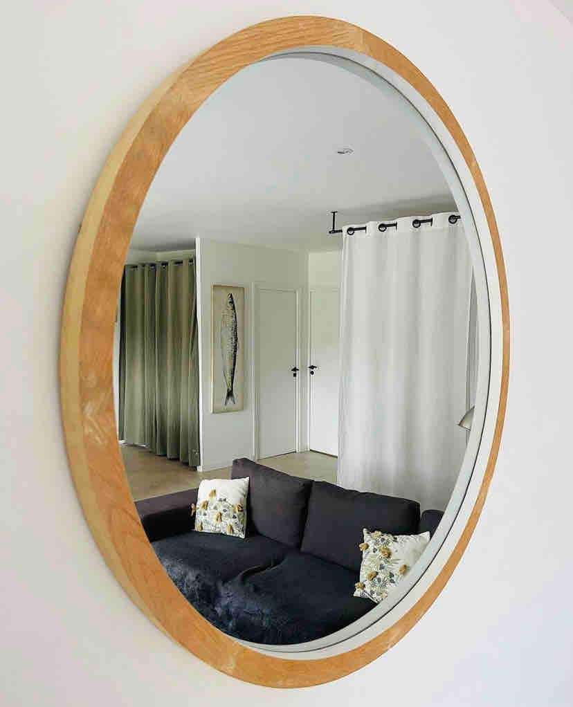 An oval mirror with a wooden frame reflects a cozy living area, including a dark sofa adorned with decorative cushions. Soft light filters through nearby curtains, enhancing the space's tranquil ambiance.