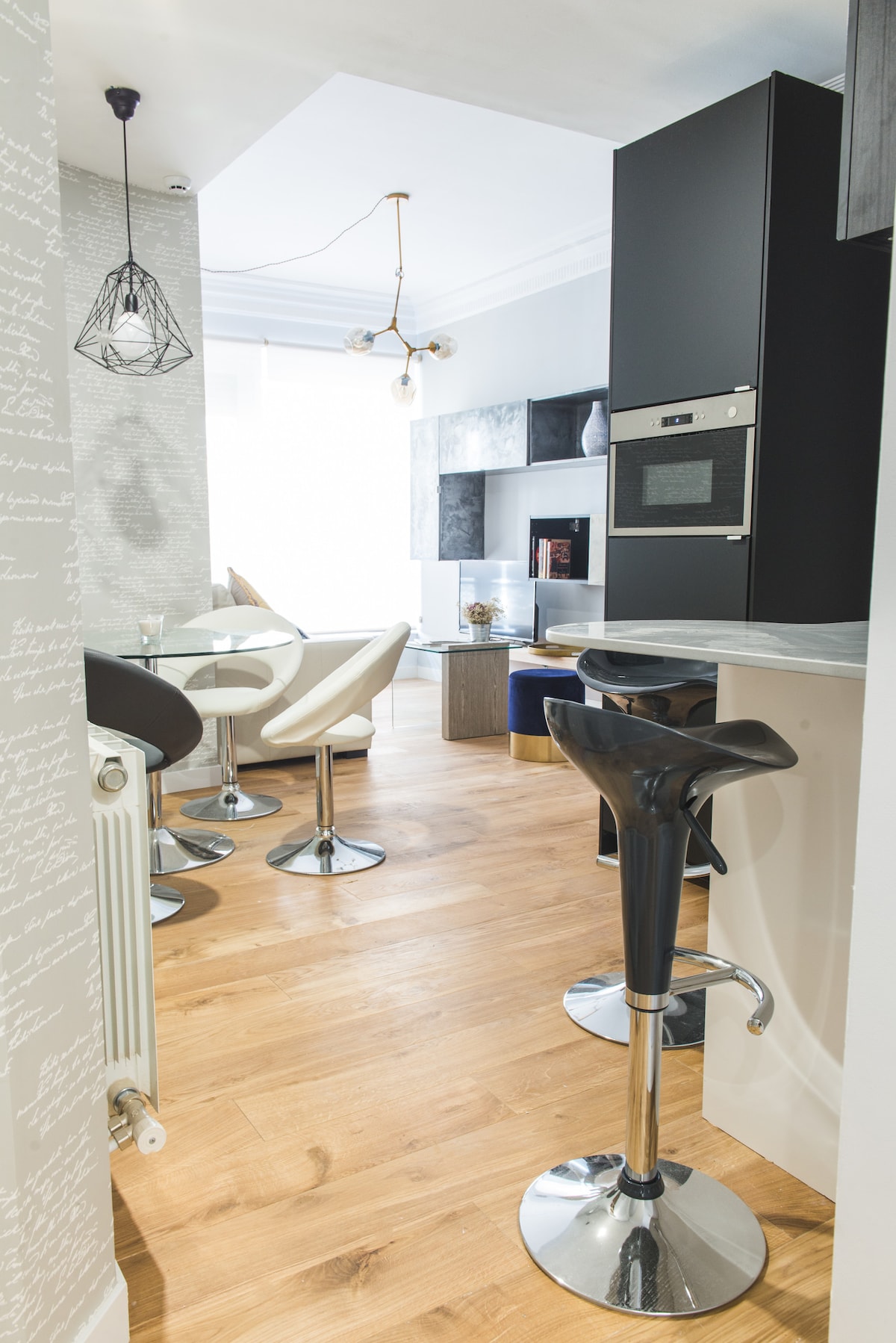 A modern kitchen and dining area is displayed, featuring sleek bar stools and a circular dining table. Light oak flooring enhances the space, which is illuminated by natural light coming through large windows. Contemporary cabinetry and an oven are visible in the nearby kitchen area.