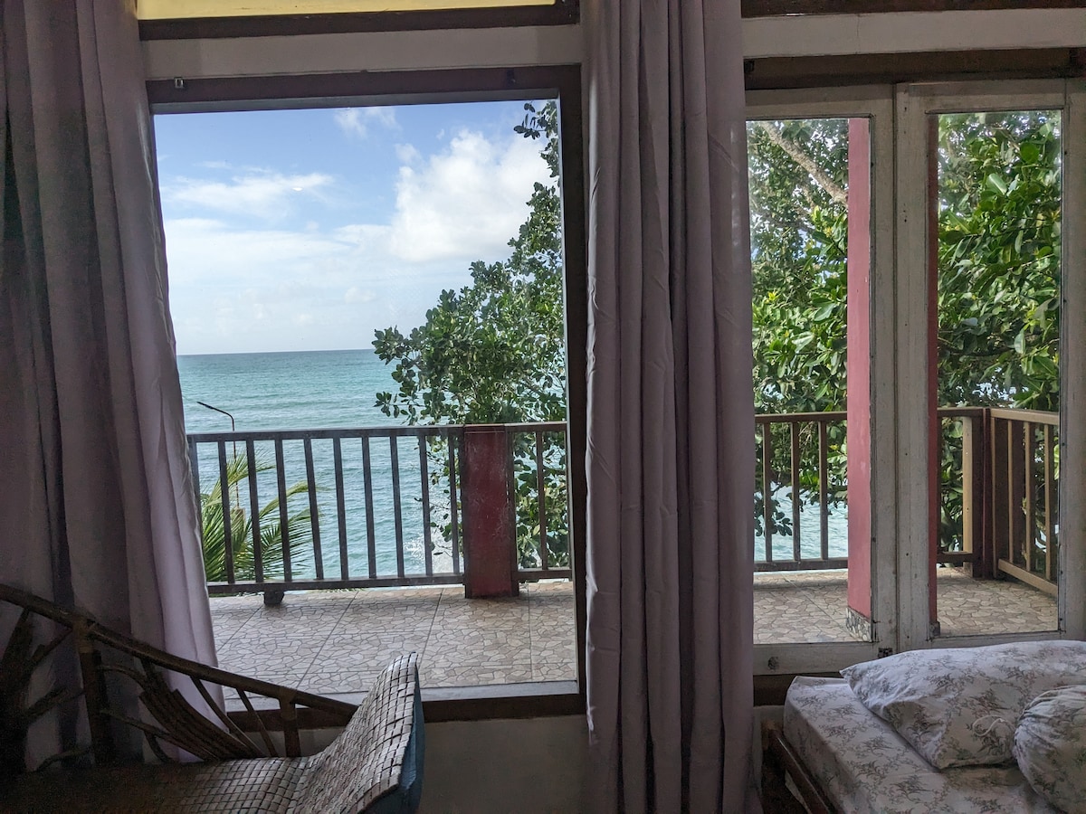 A view from an interior room showcases large glass doors leading to a private balcony. The serene ocean is visible through the open space, framed by lush greenery. Light curtains gently sway, allowing natural light to illuminate the area.