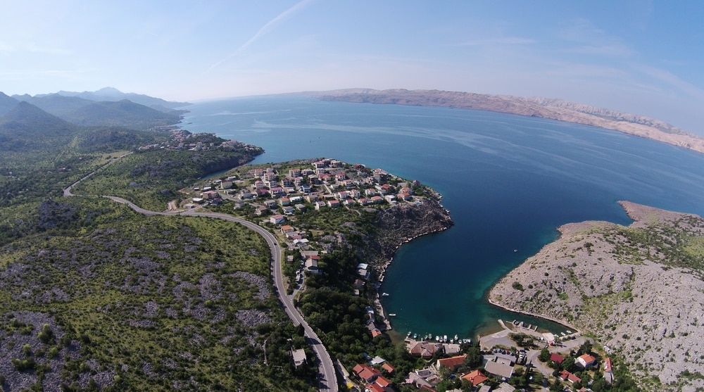 An aerial view captures the scenic coastline near Cesarica, showcasing lush green hills and tranquil blue waters. The layout of nearby residential areas is visible, along with a winding road. In the distance, the outline of a mountainous landscape and an expansive fjord can be observed.