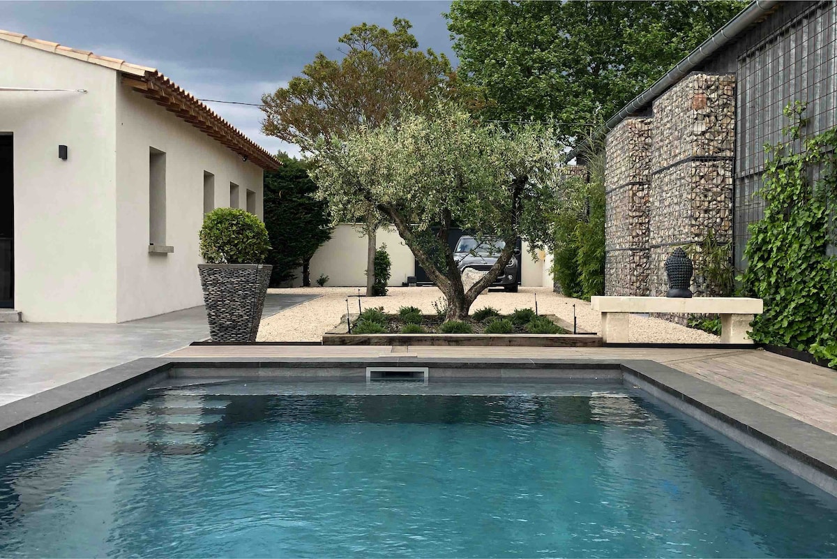 A private pool is visible in the foreground, surrounded by a wooden deck. In the background, landscaped gardens include an olive tree and various shrubs, with a stone wall partially obscured by greenery. The sky features a soft gray hue, adding to the serene atmosphere.
