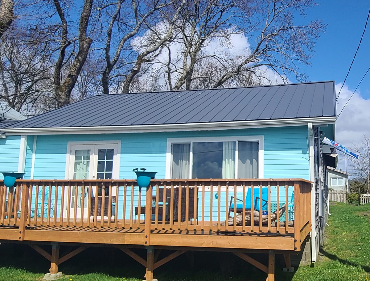 A light blue house is featured alongside a wooden deck, which is equipped with lounge chairs and decorative planters. French doors provide access to the interior, while the backdrop showcases a clear blue sky and trees. The setting offers a serene environment.