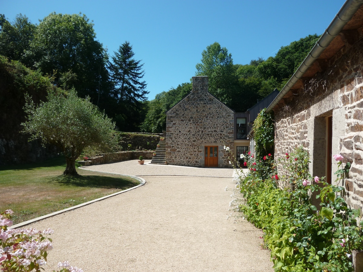 A charming stone building is set against a clear blue sky, surrounded by well-maintained gardens. A gravel path leads to the entrance, bordered by flowering plants and greenery. Trees provide a natural backdrop, enhancing the serene outdoor space.