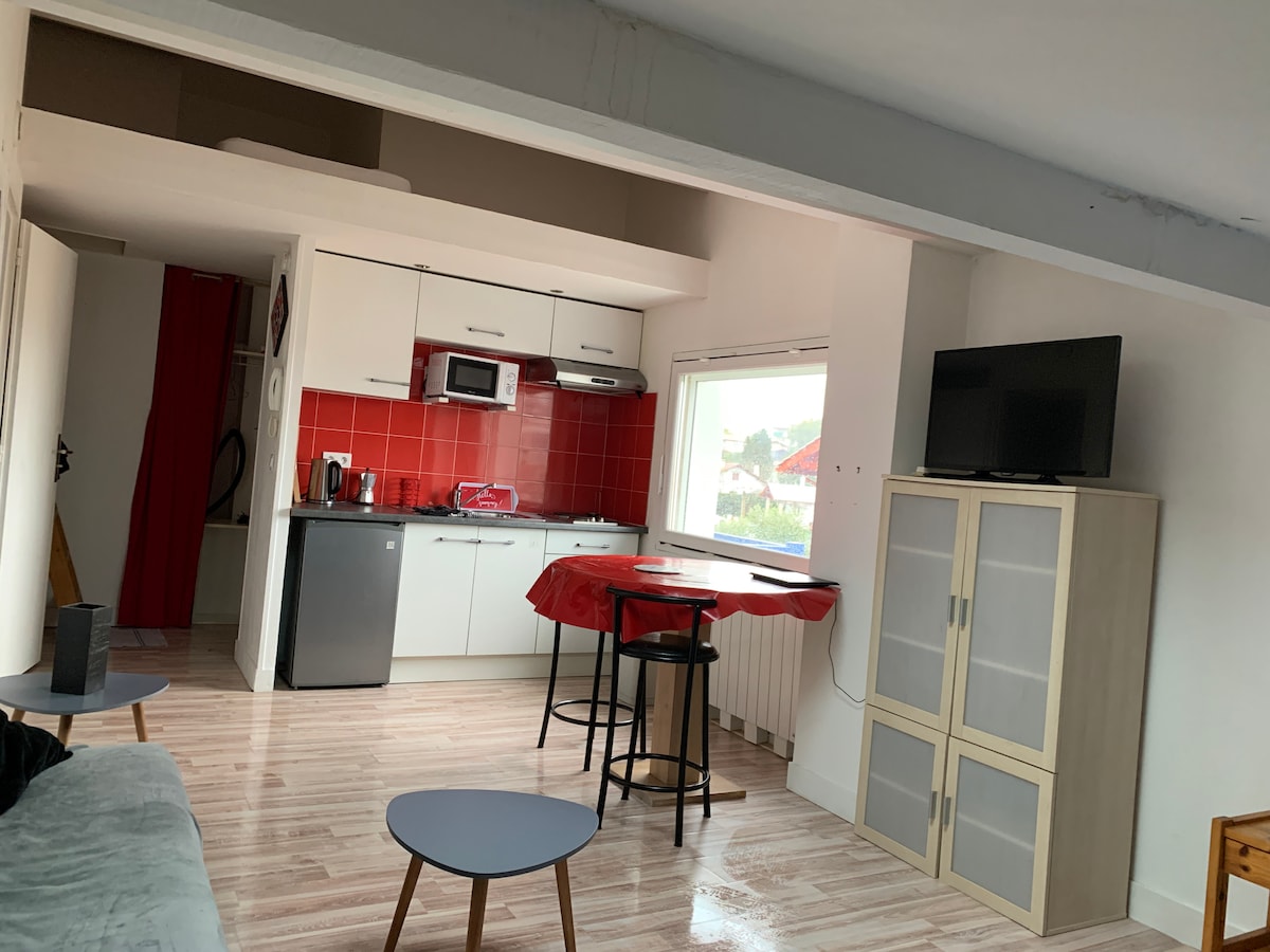 A bright studio space is displayed, featuring a compact kitchen with white cabinets and red tile accents. A dining table set for two is placed near a window, alongside a small flat-screen TV mounted on a light-colored cabinet. The floor is covered in light wooden planks, enhancing the airy feel.