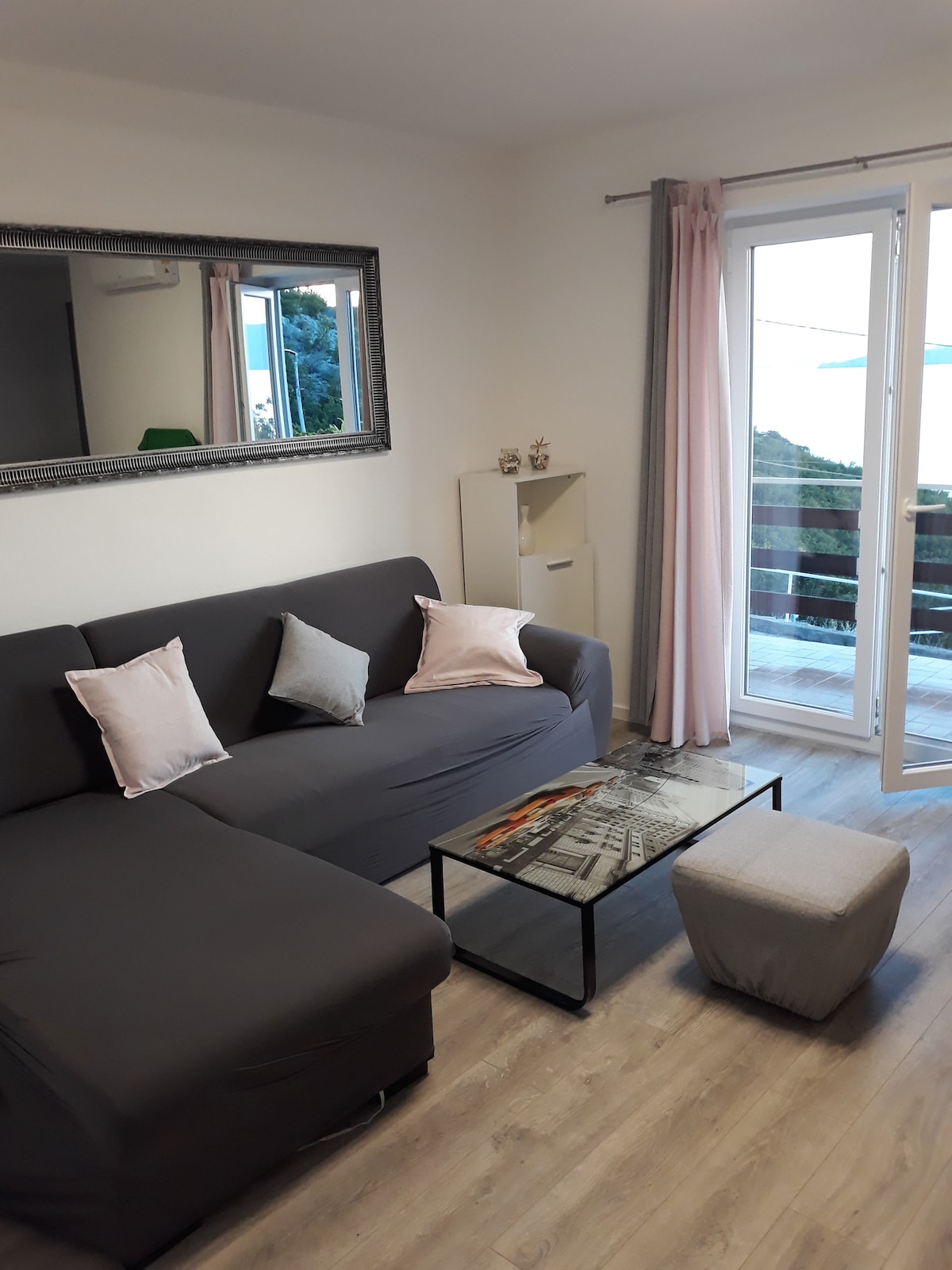 A cozy living area is highlighted by a sectional sofa adorned with soft pillows. A glass coffee table rests in front, complemented by a cushioned ottoman. Large glass doors offer access to a balcony with views of the surroundings, allowing natural light to fill the space.