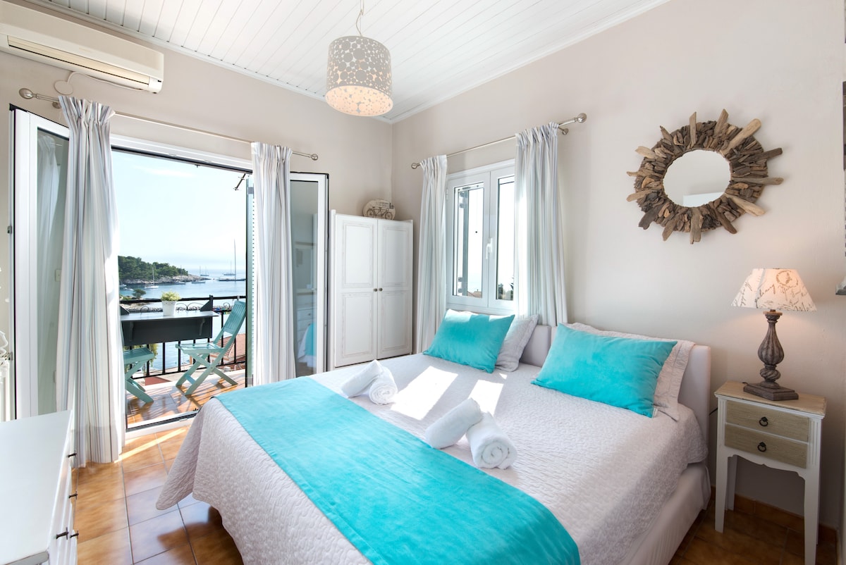 The bedroom features two single beds, each adorned with soft turquoise bed runners. Large windows open to a balcony, revealing a view of the surrounding landscape. A mirror with a natural design is mounted on the wall, and a decorative lamp provides gentle lighting.