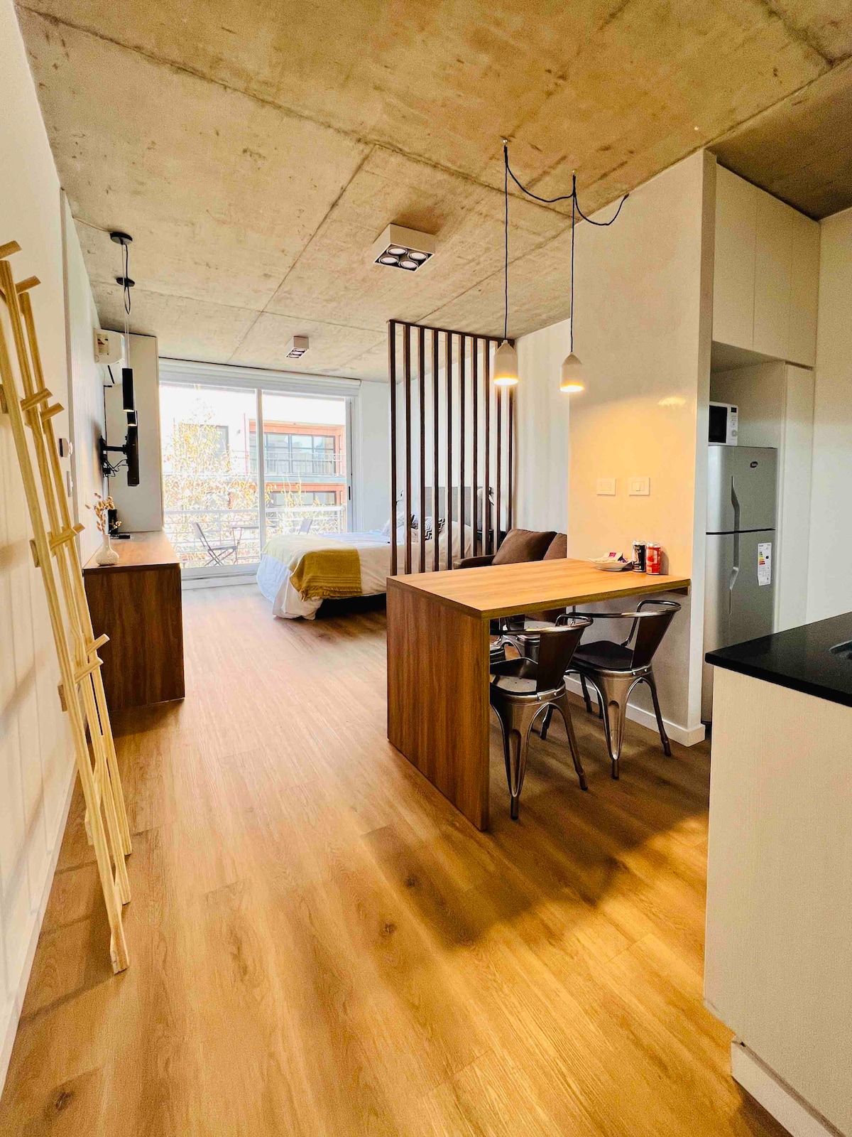 An open-concept living space features a wooden dining table with two chairs and a kitchenette equipped with modern appliances. Light floods in from large windows revealing a cozy sleeping area in the background, separated by a stylish wooden partition.