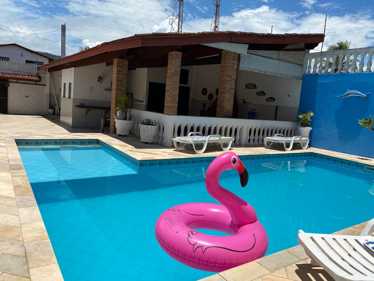A spacious swimming pool is framed by a tiled deck, featuring a large pink inflatable flamingo. In the background, an outdoor entertainment area is visible with a rustic covered space, complemented by various seating options and a blue wall.