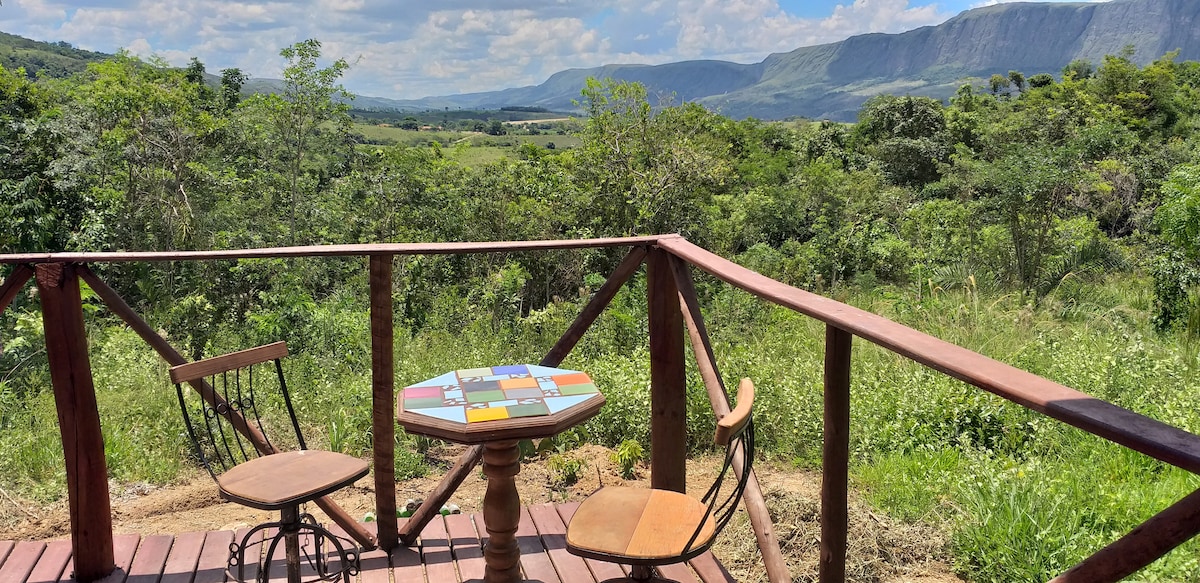 A wooden deck is shown with a colorful round table and two chairs, overlooking a lush green landscape. Rolling hills and distant mountains are visible under a partly cloudy sky, creating a serene outdoor space for relaxation.