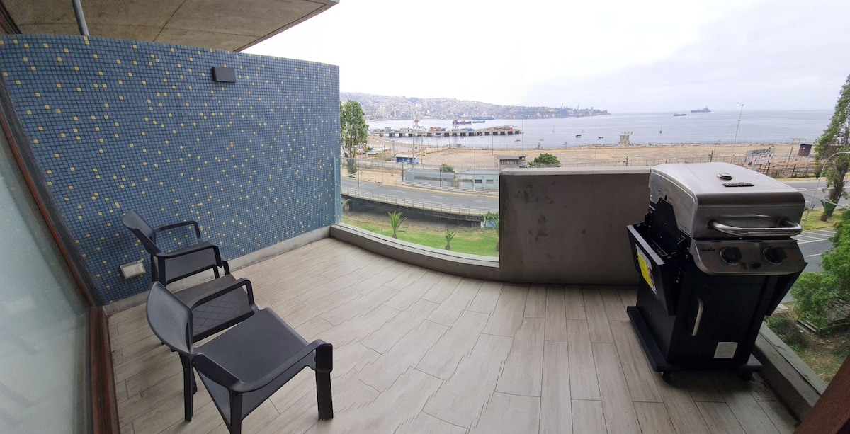 An outdoor terrace features two black lounge chairs positioned for ocean views. A barbecue grill is located nearby, and the scenery includes sandy beaches and coastal water. The backdrop shows a city landscape and harbor, creating a serene atmosphere.