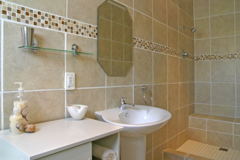 A well-appointed bathroom is featured, showcasing tiled walls in soft earth tones. A modern sink is situated beside a glass shower stall. A small shelf displays toiletries, while a decorative bowl adds a touch of elegance. Soft lighting enhances the serene ambiance.