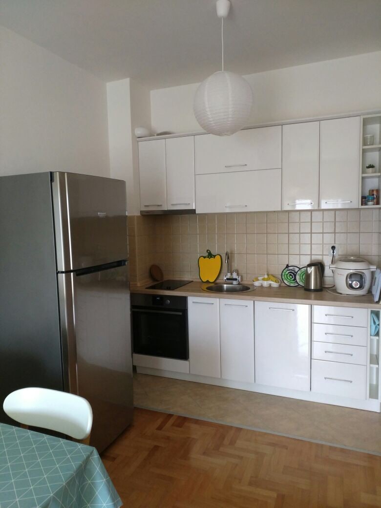 A modern kitchen is displayed with sleek white cabinetry and a stainless steel refrigerator. A countertop includes an electric kettle and a rice cooker. A dining table with a patterned tablecloth is present in the foreground, enhancing the kitchen's functionality.