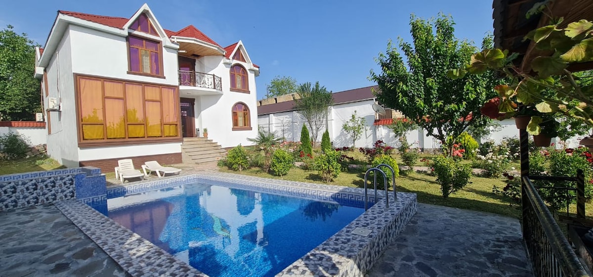 A two-story house with a welcoming facade is surrounded by a well-maintained garden. A clear blue pool reflects the sky, with lounge chairs positioned neatly beside it. Lush greenery and colorful plants frame the property, enhancing the peaceful environment.